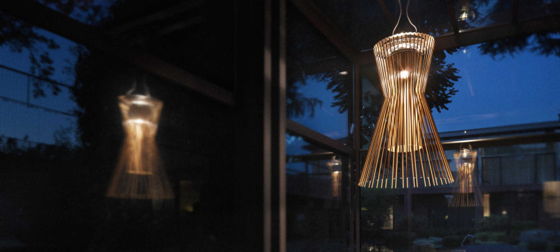Suspension Allegro Vivace de Foscarini disposée dans une pièce vitrée avec la lumière du jour qui tombe. Cette dernière est en aluminium verni et métal chromé et est de couleur cuivre. Allegro Vivace est composé de lignes géométriques essentielles et habillé de reflets cuivrés.