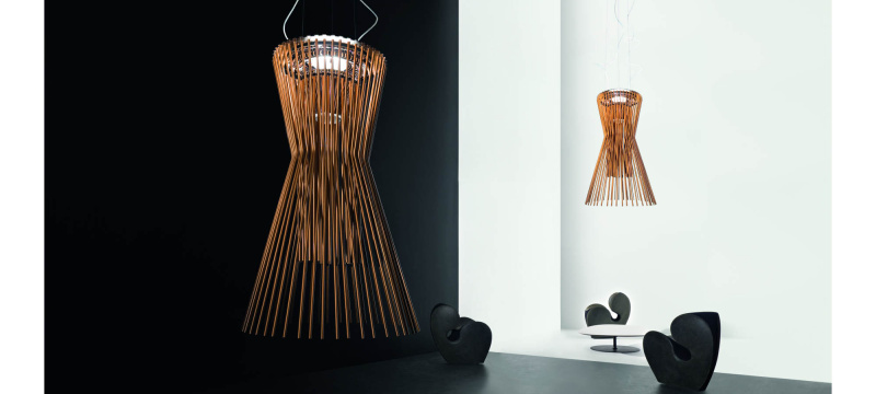 Suspension Allegro Vivace de Foscarini disposé dans une pièce épurée blanche avec un fauteuil noir. Cette dernière est en aluminium verni et métal chromé et est de couleur cuivre. Allegro Vivace est composé de lignes géométriques essentielles et habillé de reflets cuivrés.