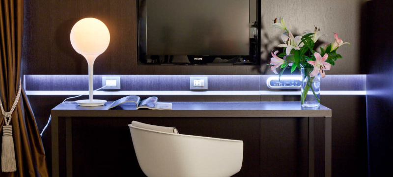 Lampe de table Castore d'Artemide disposée sur le bureau d'une chambre d'hôtel. Ce dernier dispose d'une base en zamac, d'une tige en résine thermoplastique et d'un diffuseur de lumière en verre soufflé.
